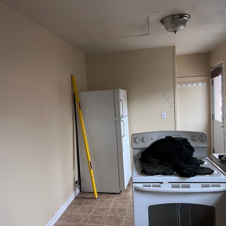 A kitchen with a refrigerator, stove, and a yellow level leaning against the wall. Beige walls, linoleum floor.