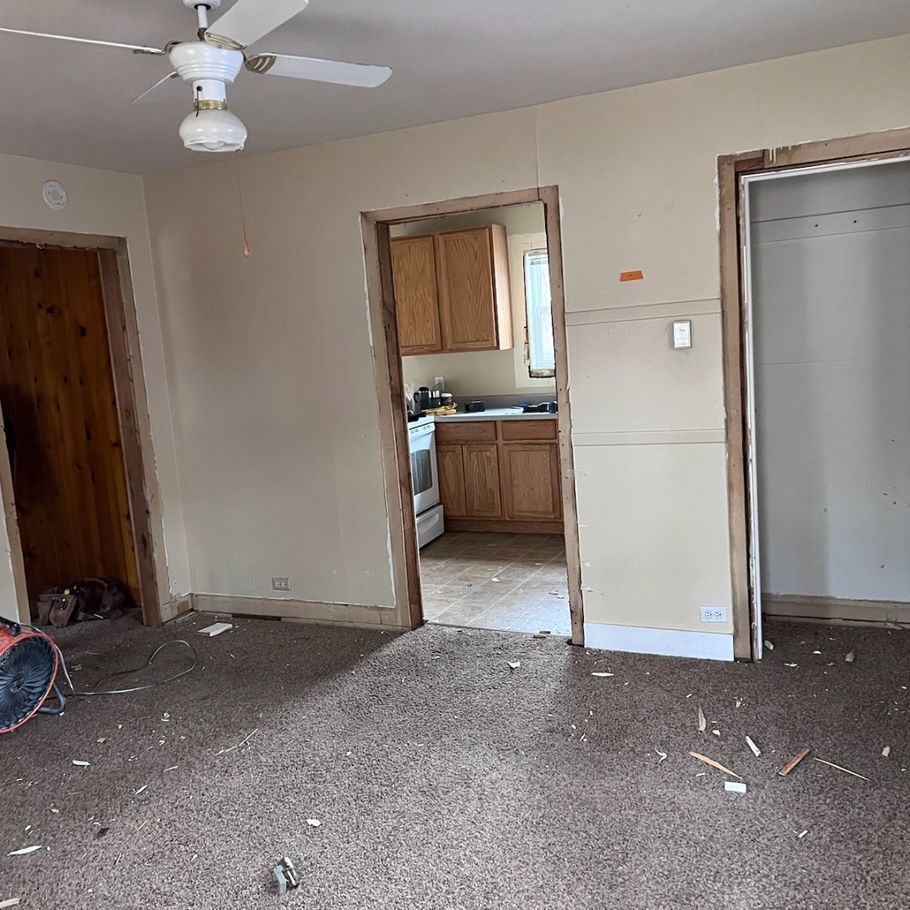 A room undergoing renovation. The beige-walled space has partially framed doorways, visible kitchen, and debris on the carpet.
