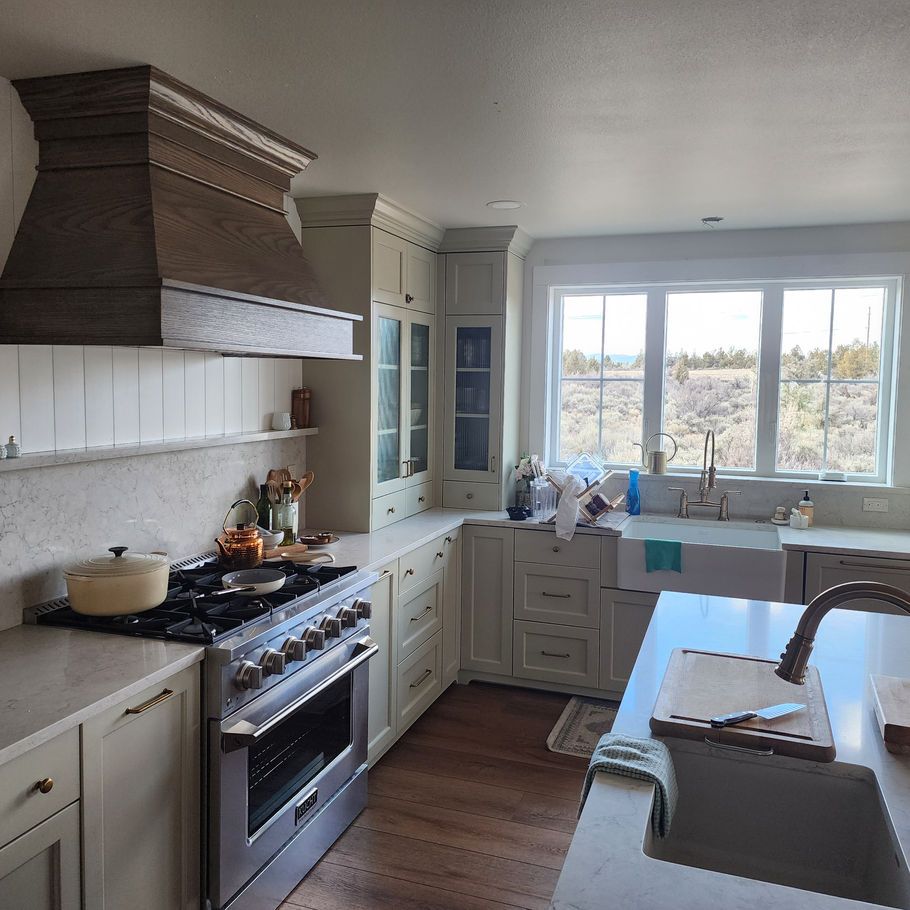 A modern kitchen with light-colored cabinets and countertops, a stainless steel oven, and a large window overlooking a natural landscape.