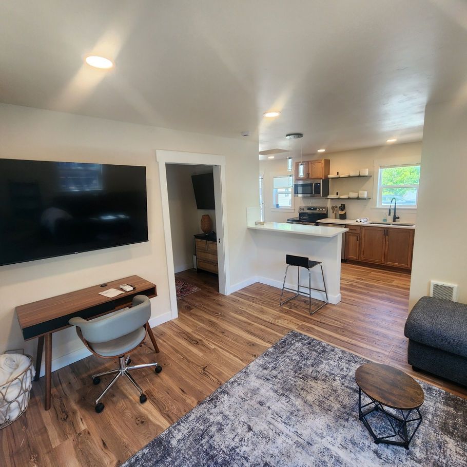 A living space with a kitchen in the background. There's a desk, a TV, a rug, and a couch. Hardwood floors throughout.