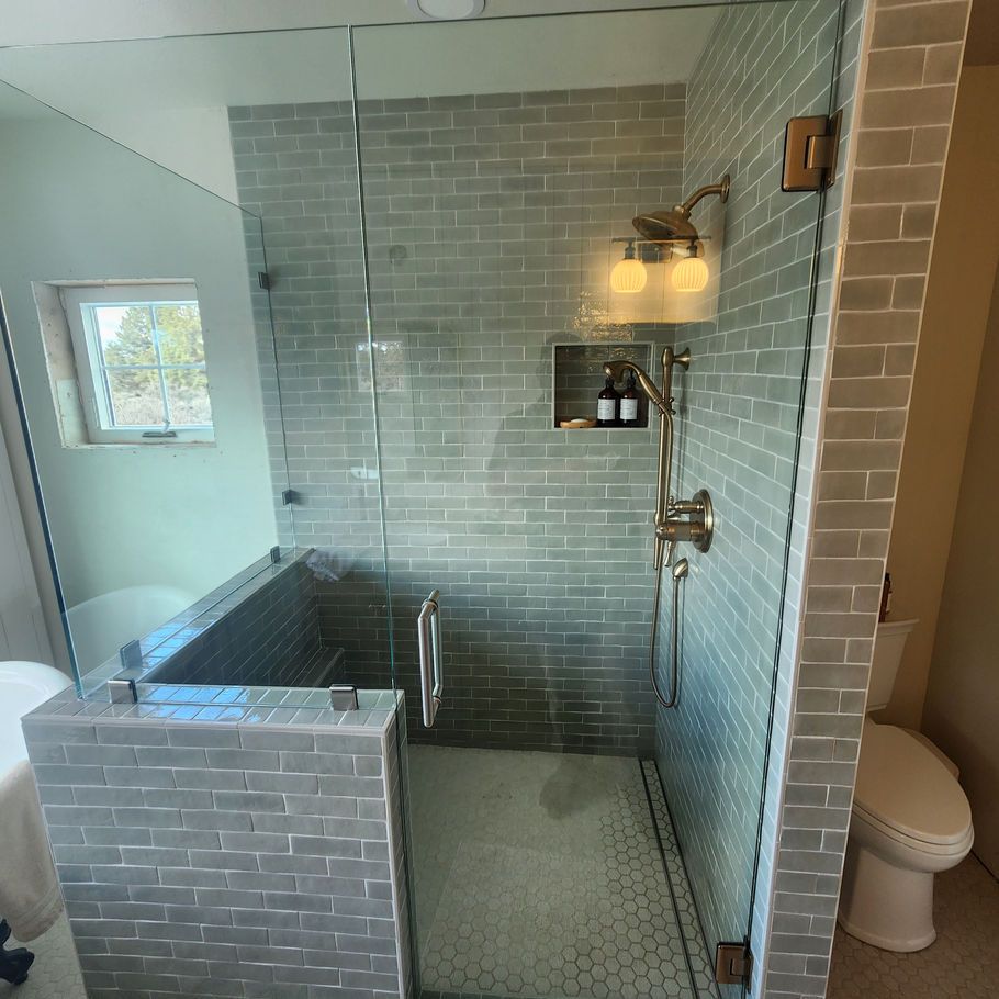 A modern shower with gray brick tiles, a glass door, and a built-in shelf. A window is visible on the left.