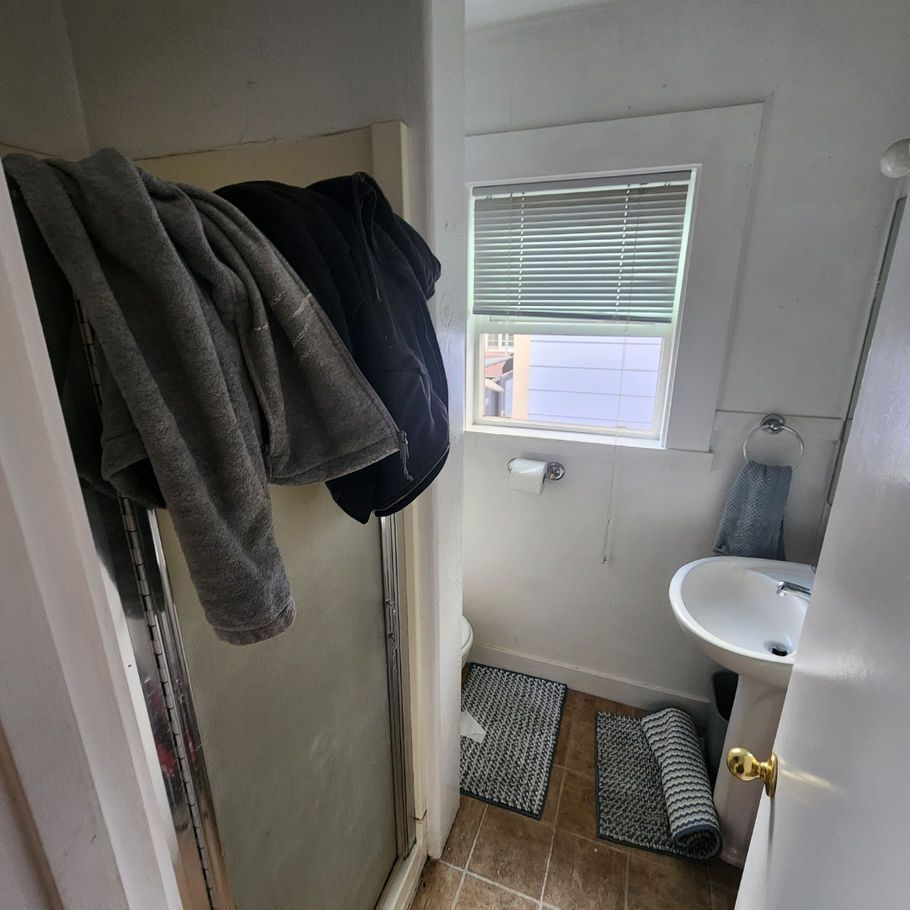A small bathroom with a shower, sink, and window. Clothing hangs inside the shower, with mats on the floor.