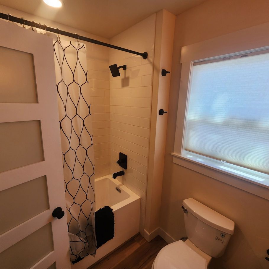 A small bathroom with a white bathtub, toilet, and window. The shower has a black showerhead and curtain.