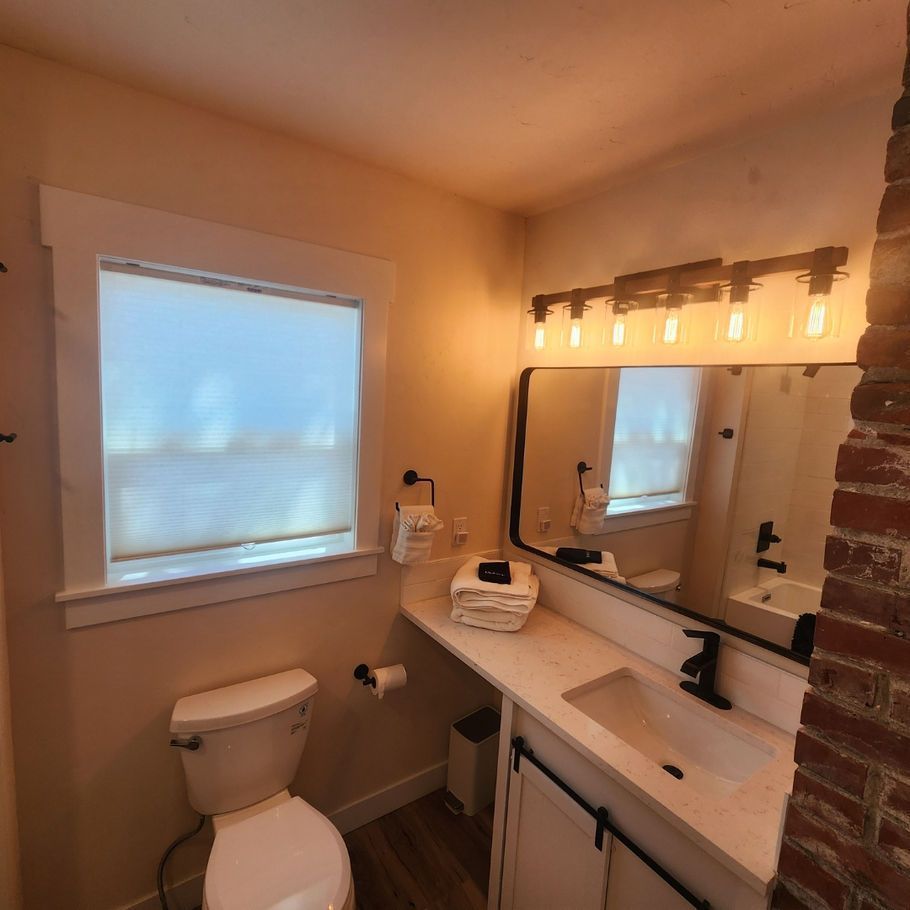 Small bathroom with a white toilet, sink, and vanity. A mirror hangs above the sink, reflecting a window and a brick wall.