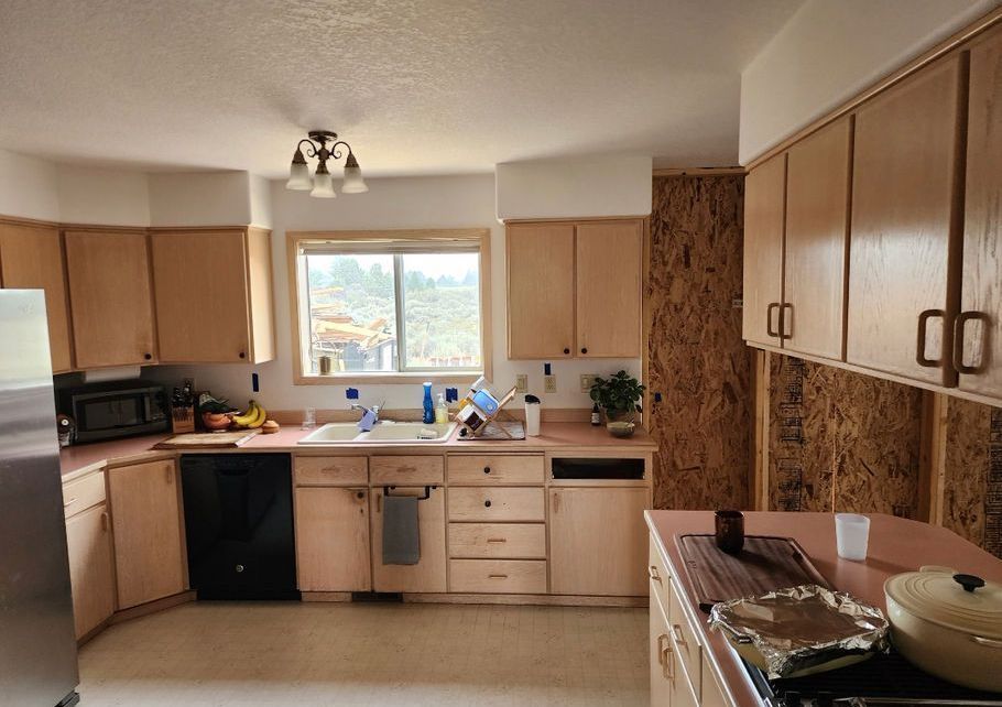 Kitchen undergoing renovation with exposed wooden wall, light cabinets, black appliances, and window view.