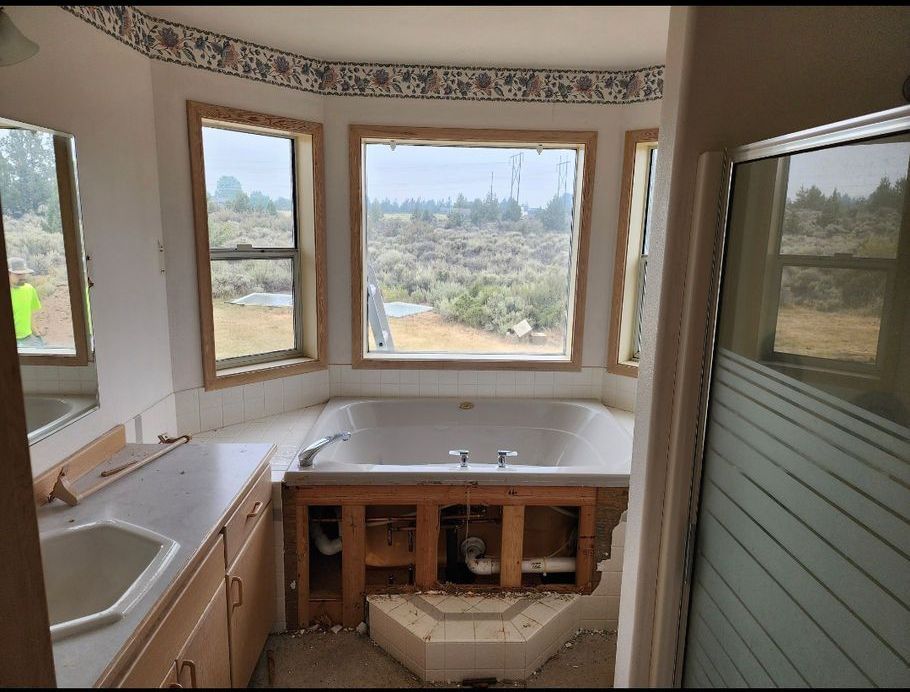 Bathroom undergoing renovation: tub surround partially removed. Beige and brown hues dominate, with windows overlooking a dry, grassy landscape.