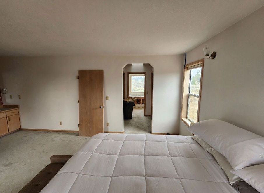 Bedroom with a bed, a door, and an arched doorway leading to another room with a window; beige walls, and carpet.