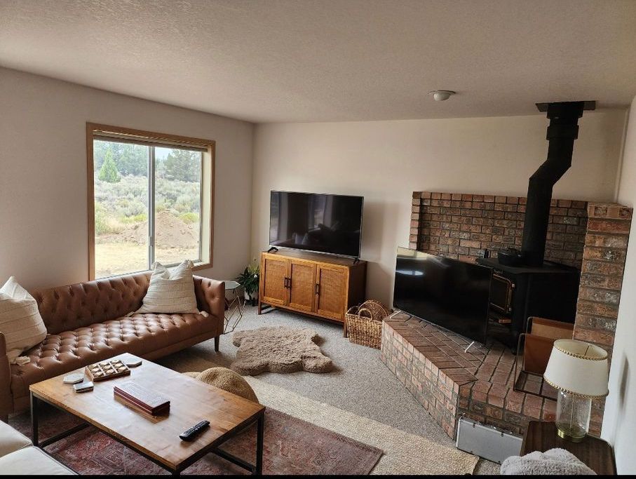 Living room with brown leather sofa, wood-paneled cabinet and TV, brick fireplace, and large window overlooking nature.