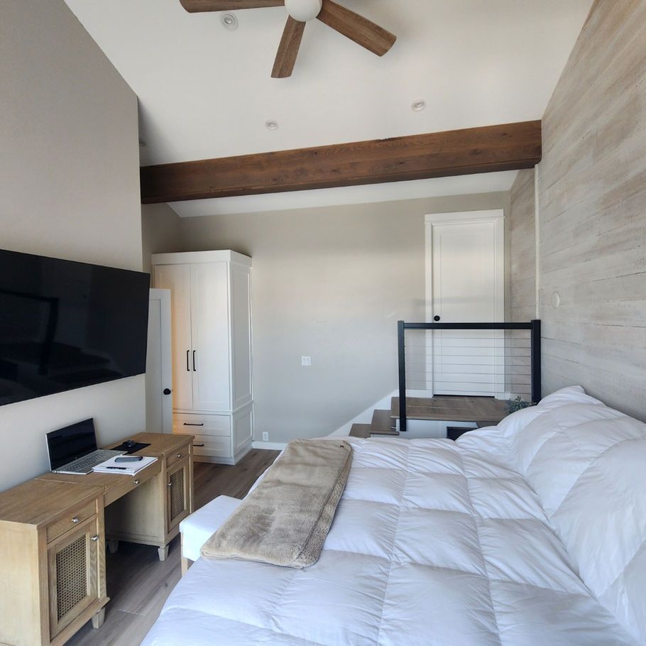 Bedroom with a white comforter on the bed, a wooden desk, and a large TV on the wall. A staircase leads to another area.