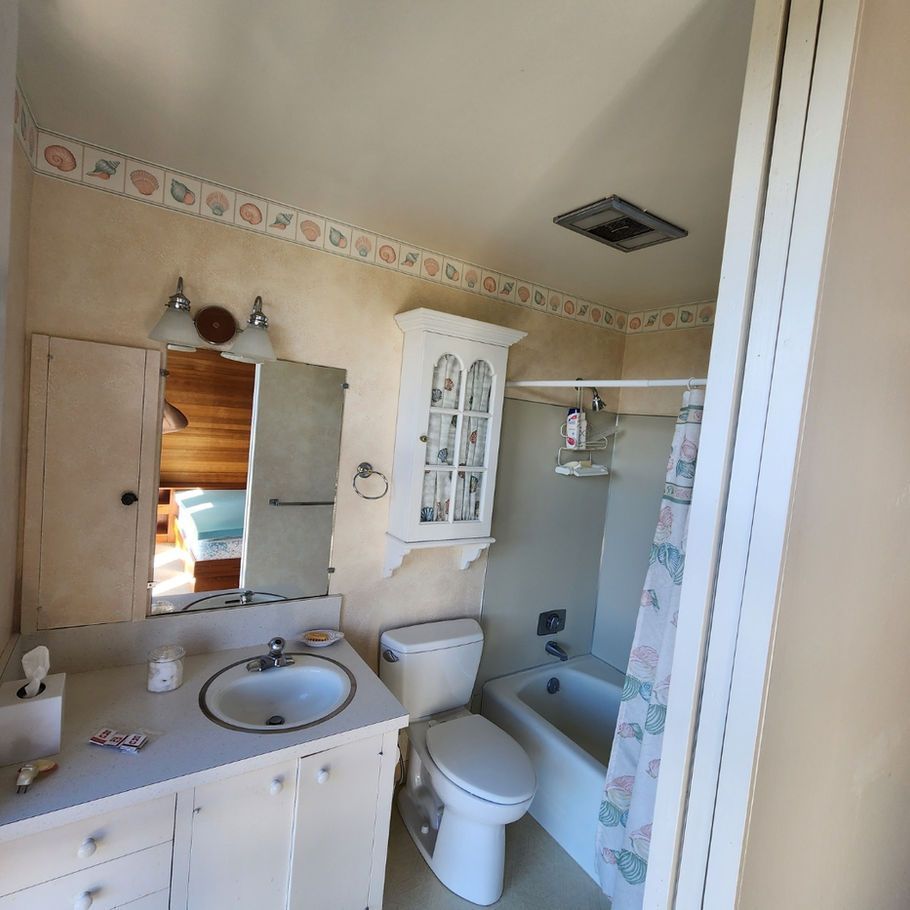 A bathroom with white cabinets and toilet, a shower/tub, and a seashell-patterned border. A mirror reflects a bedroom.