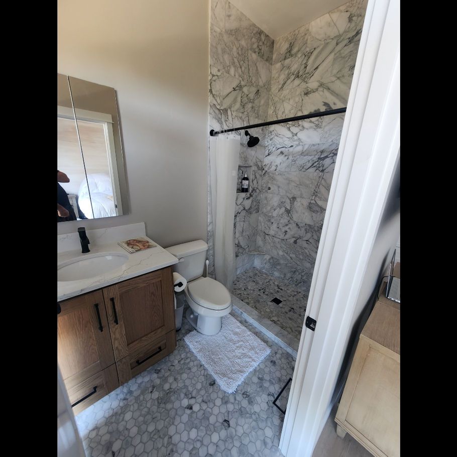 Bathroom with marble shower, wooden vanity, white toilet, and patterned floor tiles. The walls are light gray.