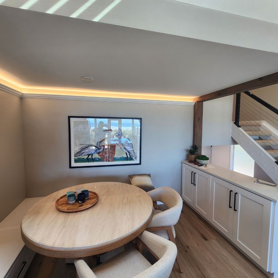 A dining nook with a round wooden table, built-in seating, and white cabinets. Artwork and accent lighting are also present.