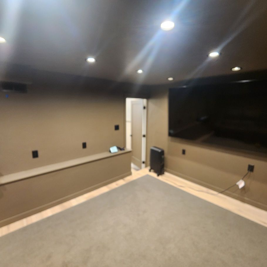 A home theater room with a large TV on the wall, a gray carpet, and a dark brown ceiling and walls. A door and a space heater are visible.