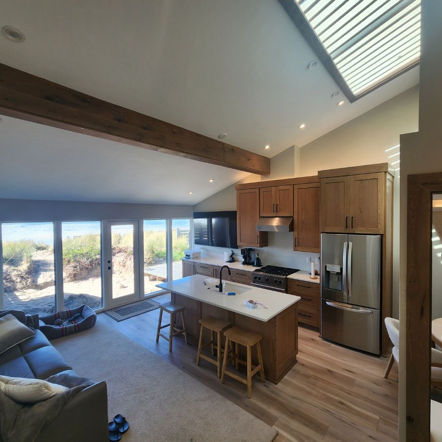 Open-concept kitchen and living space with wooden beams, a skylight, and a view of the outdoors. Features a kitchen island with stools and stainless steel appliances.