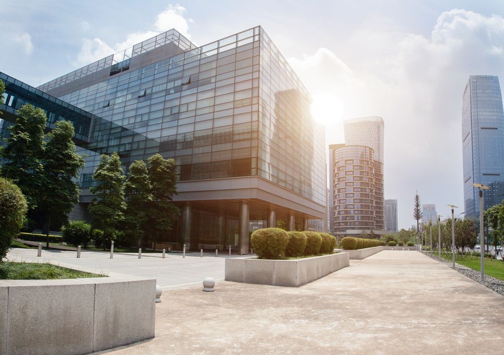 Modern glass office buildings with sunlight casting a glow on a concrete pathway lined with green shrubs and trees.