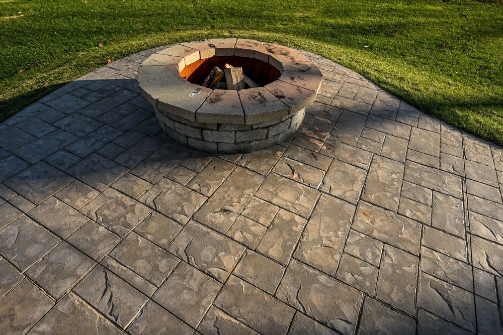 Stone fire pit with burning logs on patterned patio, bordered by green grass.