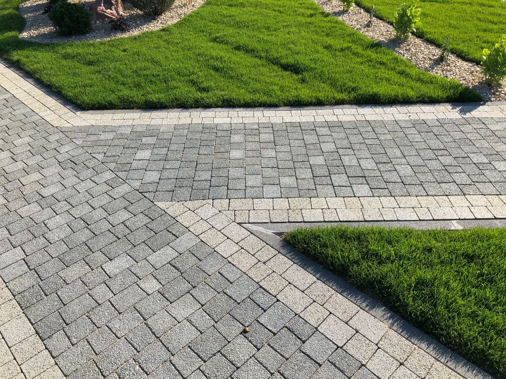 Stone paved driveway with grass accents, bordering a grassy area with young plants.