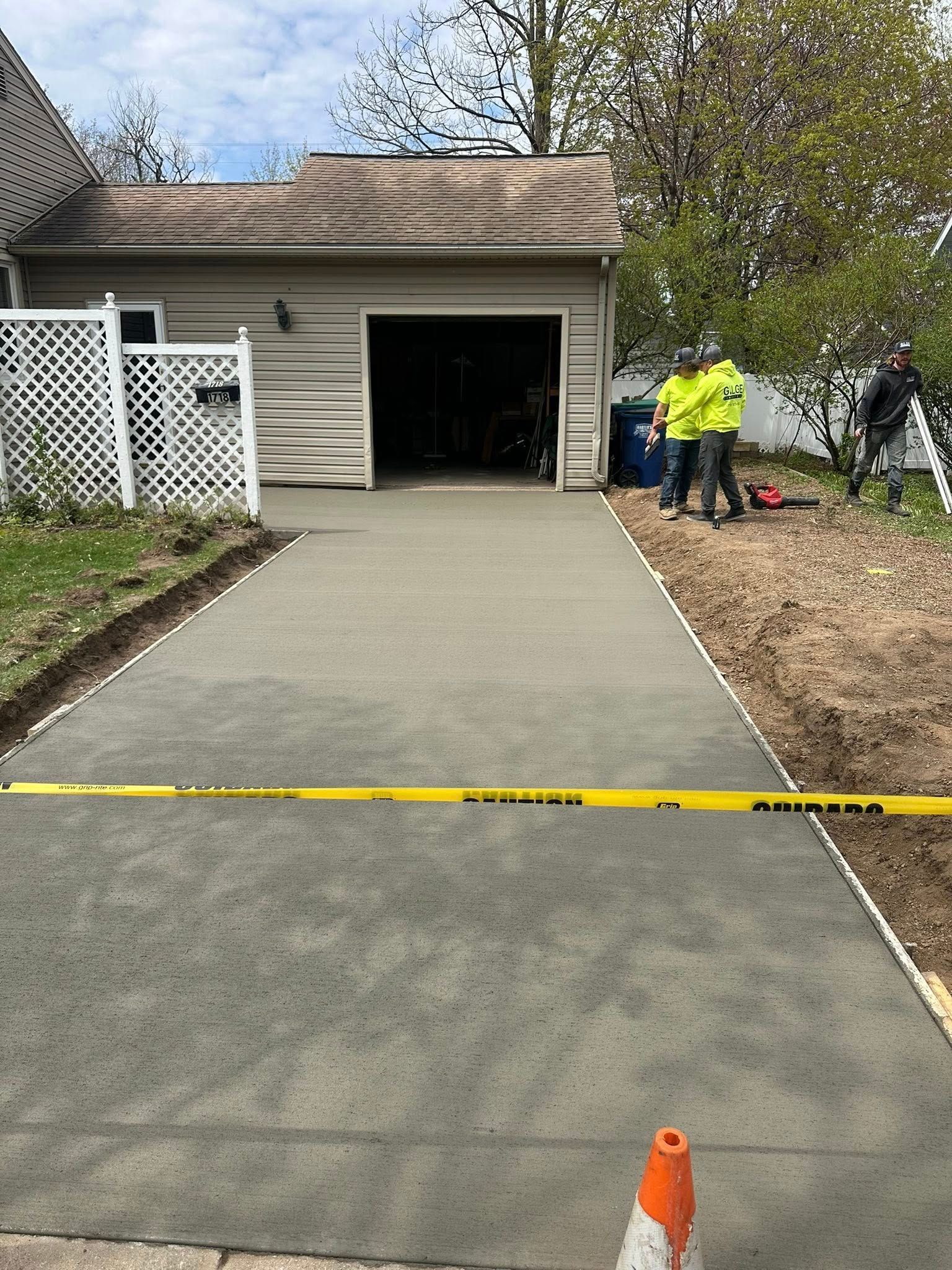 New concrete driveway leading to a garage; two workers in neon green vests, orange cone, yellow tape.