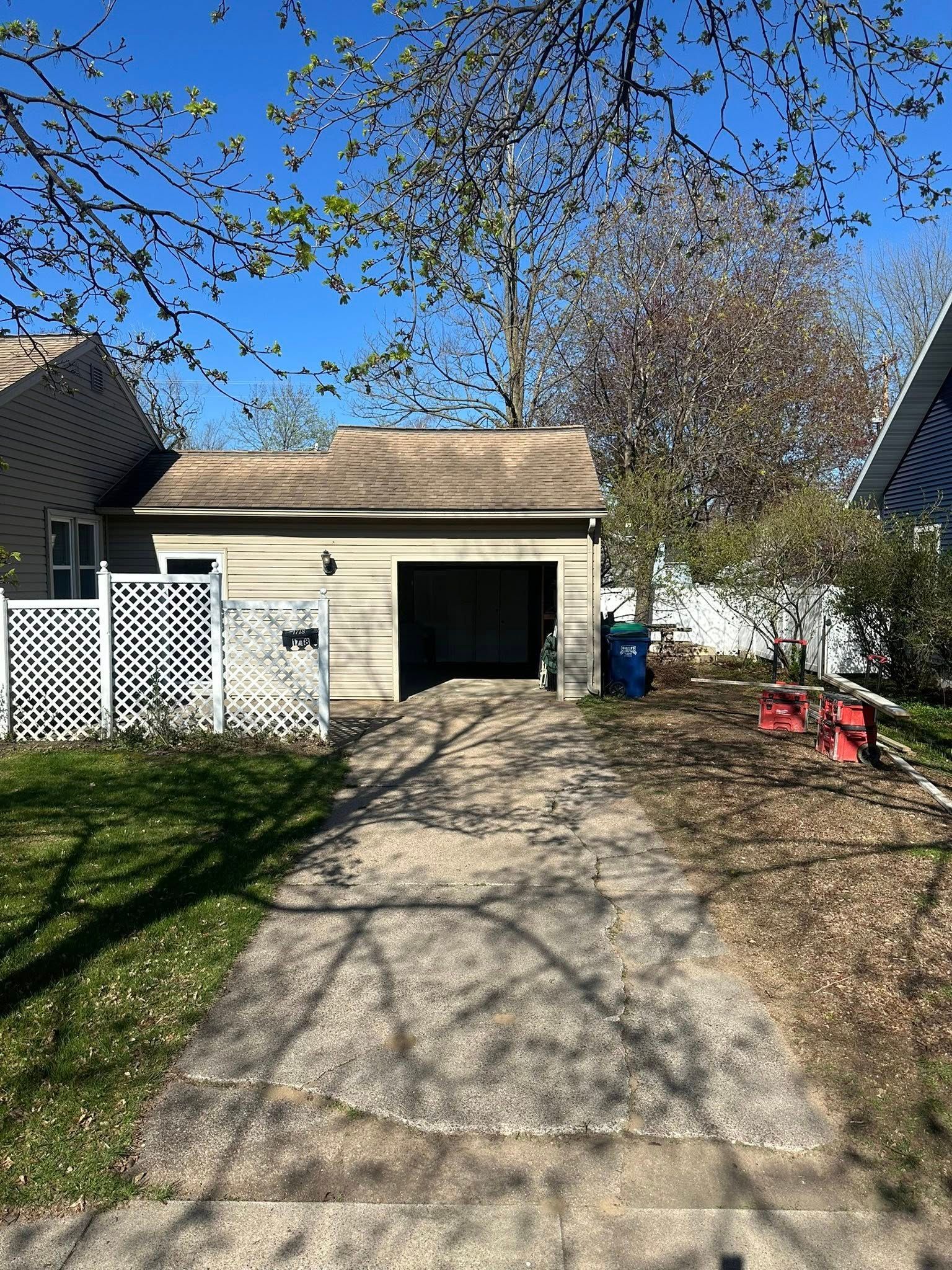 Driveway leading to a tan garage with an open door. White fence on the left, blue sky.