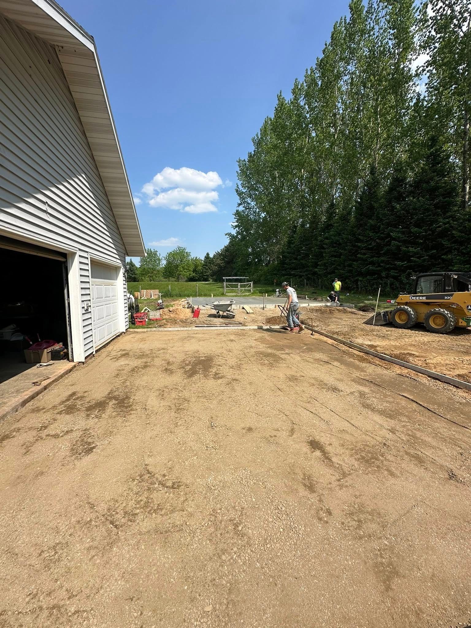 Construction site near a white garage. A person works with equipment. Gravel driveway, green trees, and blue sky.