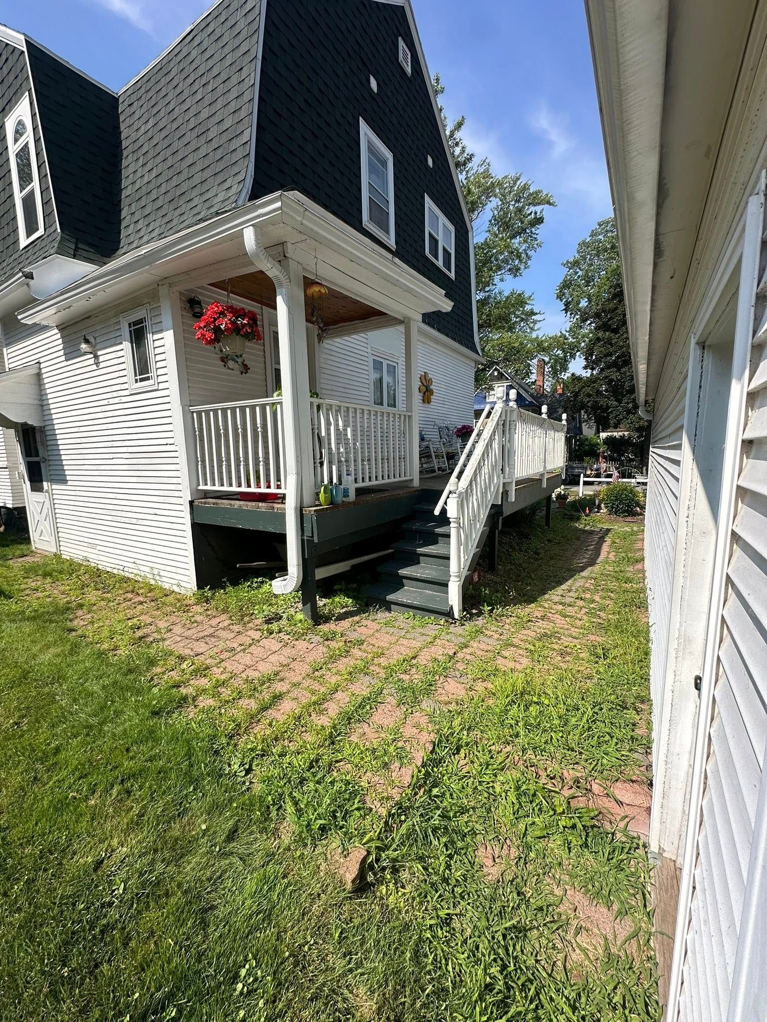Black and white house with a porch and steps, next to a white sided building on a grassy area under a blue sky.