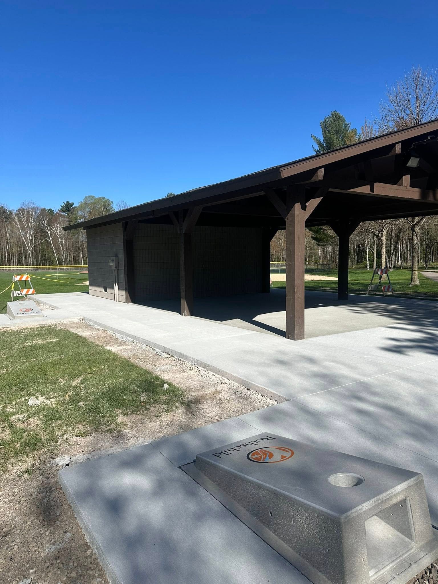 Park shelter with brown wooden beams, concrete patio, and a stone fire pit under a clear blue sky.