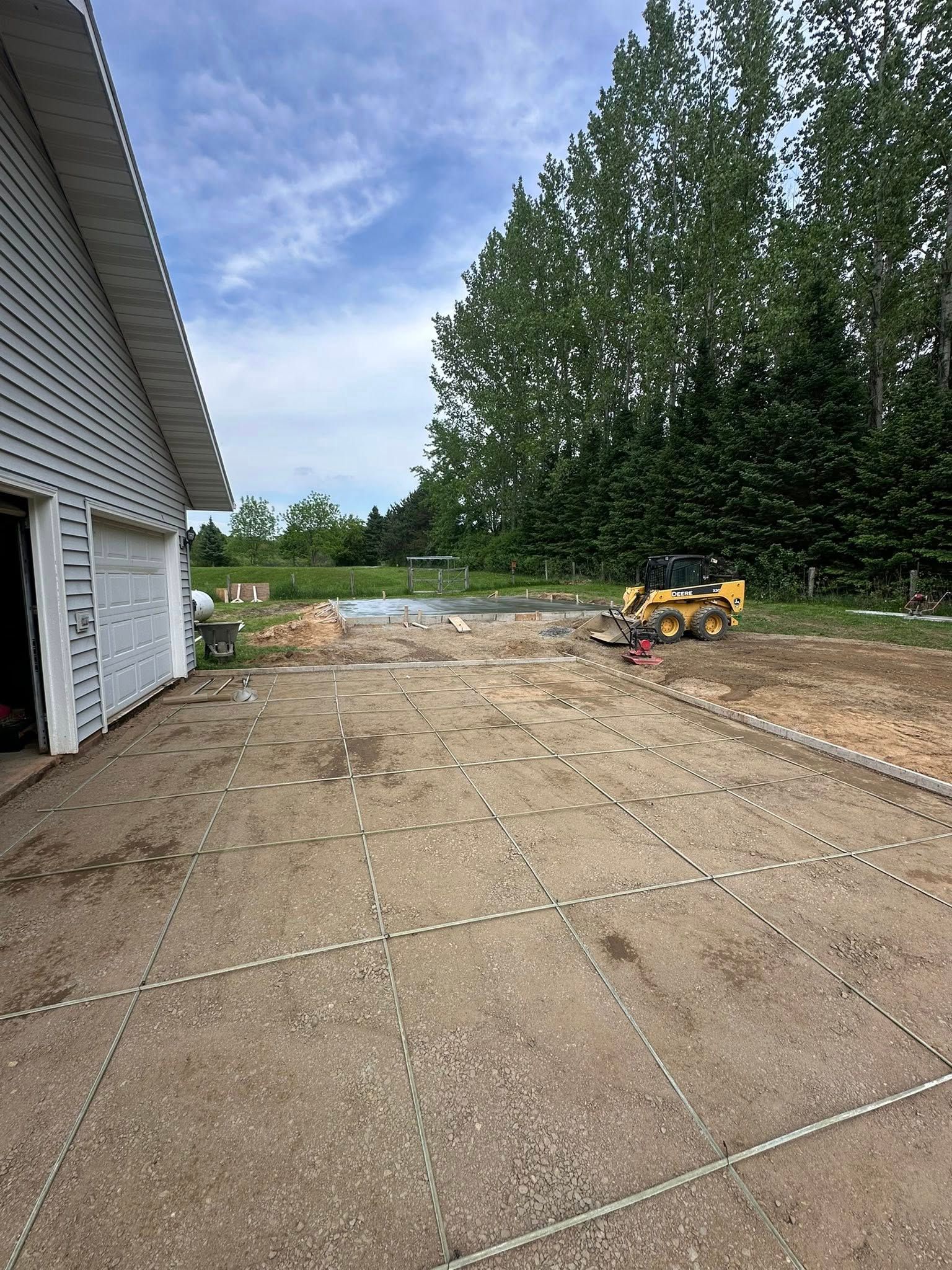 Construction site next to a garage; concrete with dotted lines, small bulldozer, trees and sky.