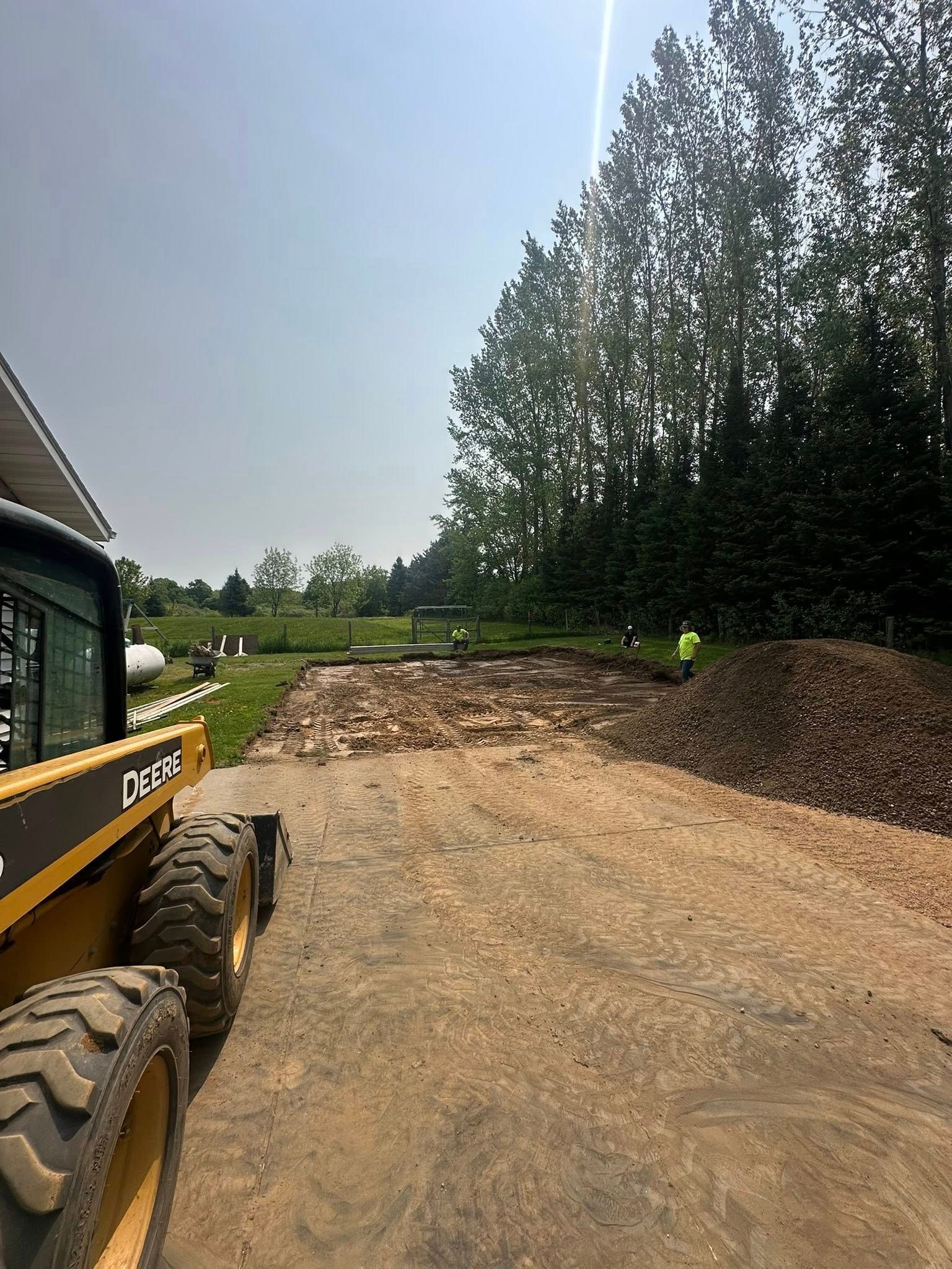 John Deere skid steer on a dirt path with a pile of mulch and trees in the background. Bright sky.