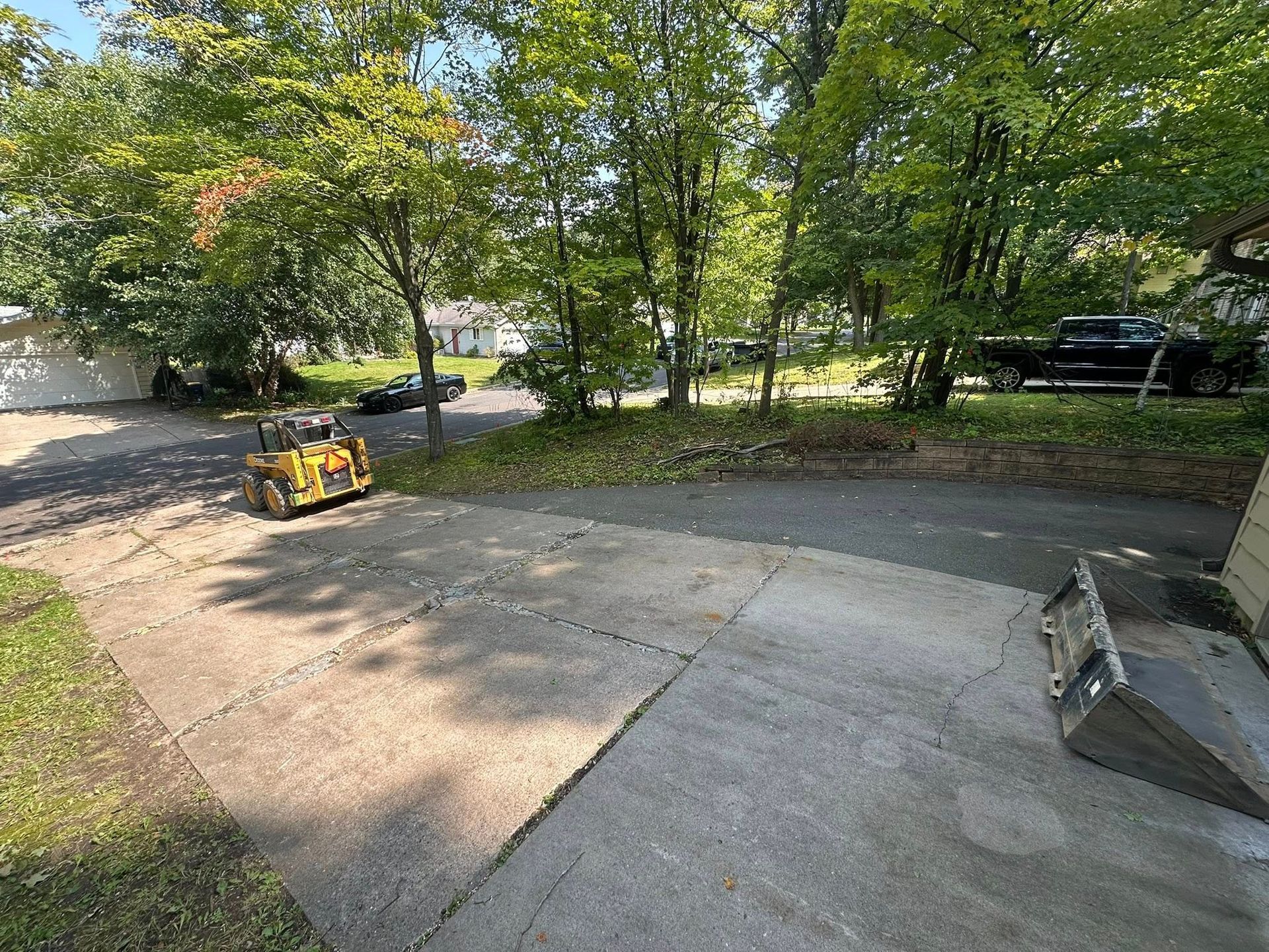 Asphalt driveway with skid steer loader, surrounded by trees and a few parked vehicles.