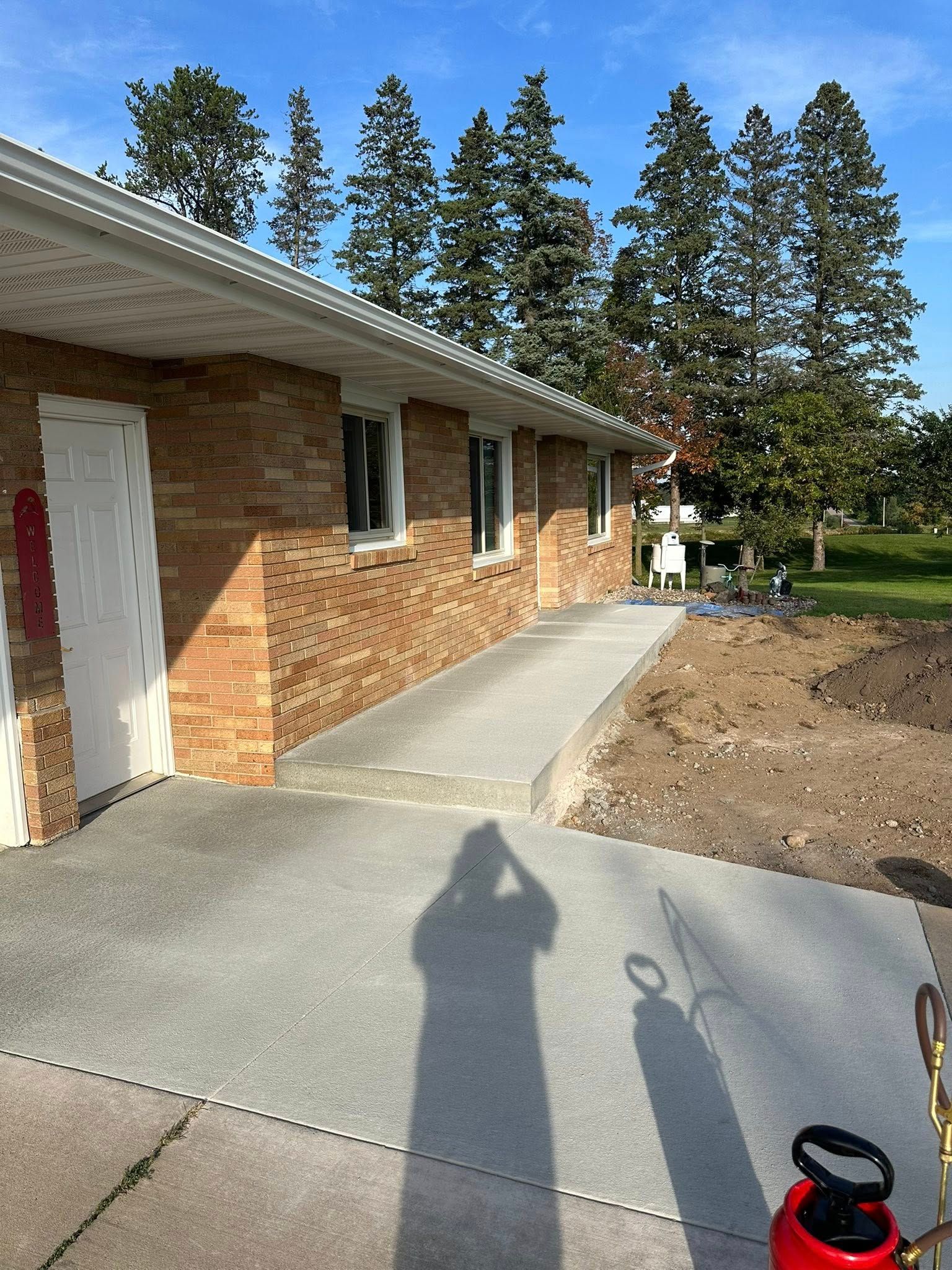 Brick building with a concrete walkway. Trees in the background. Someone's shadow in the foreground.
