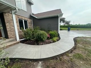 Curved concrete walkway leads to a house with brick and dark siding, surrounded by landscaping, under an overcast sky.