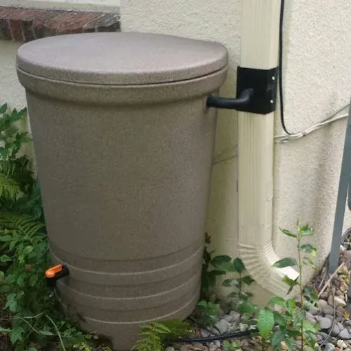 Tan rain barrel next to a downspout, with plants and a beige house wall.