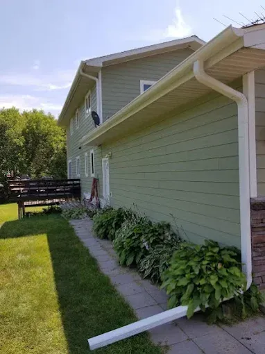 Green house with white trim, gutters, and downspout. Landscaping and a stone pathway.