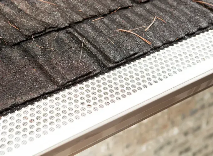 White gutter guard with small holes installed on a dark shingle roof.