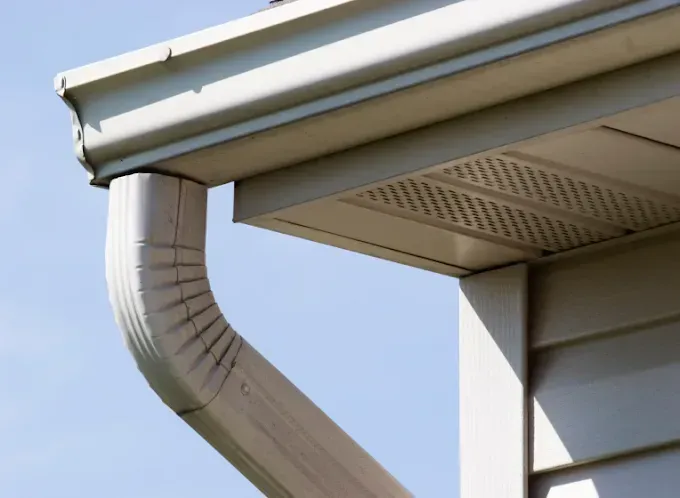 Beige gutters and downspout on the corner of a house against a blue sky.