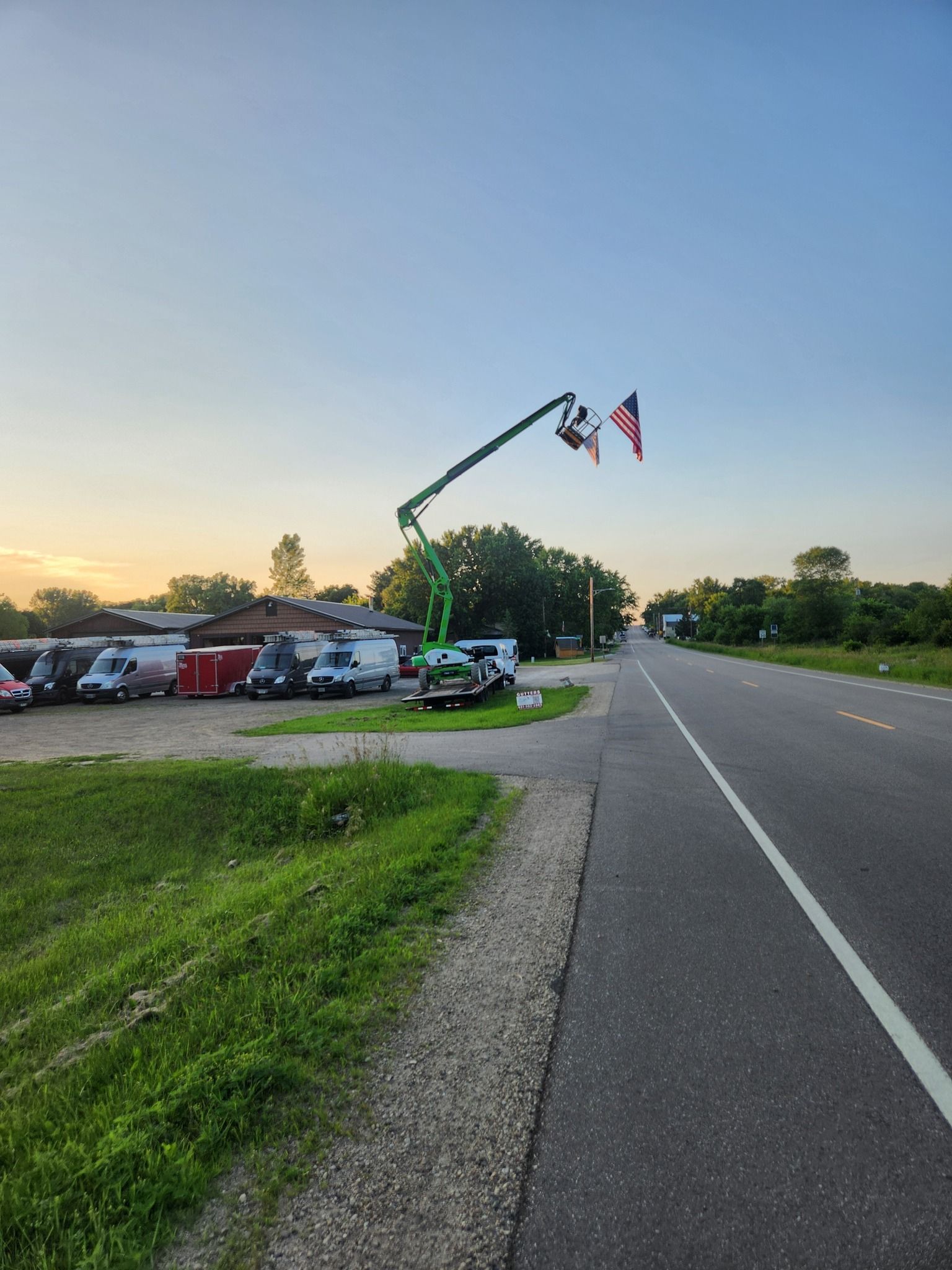A lift extends into the sky holding an American flag. It is positioned near a road, with other vehicles parked in a lot.