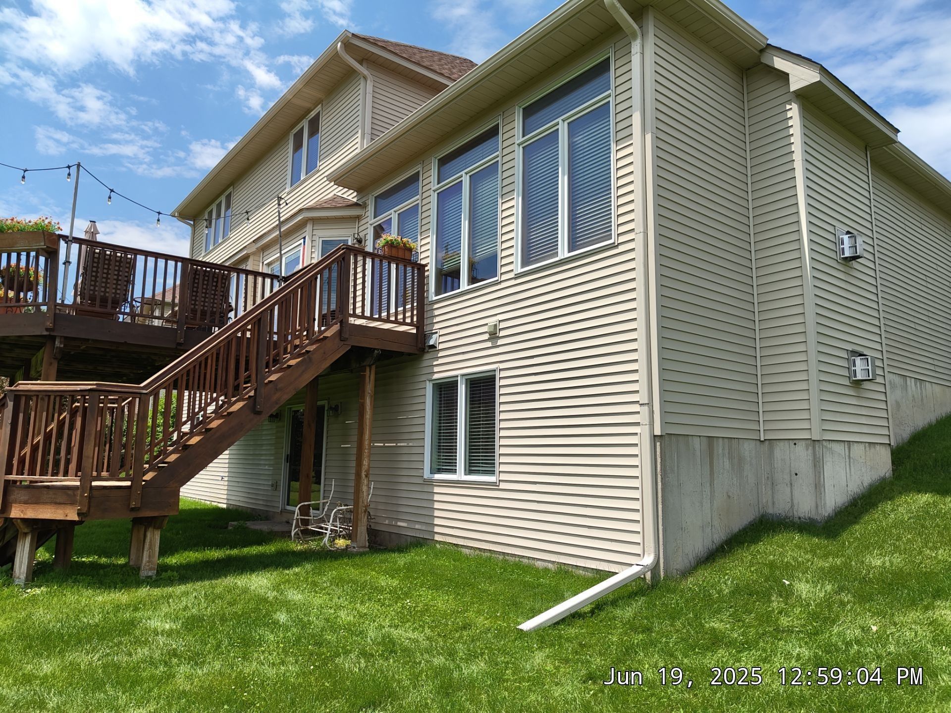 Back of a two-story beige house with a wooden deck and staircase. Green grass and blue sky are visible.
