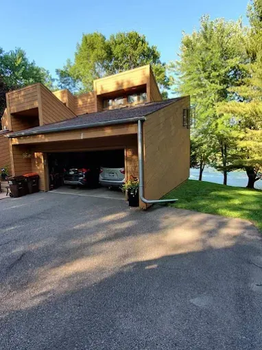 Brown house with attached garage, two cars inside, asphalt driveway, green trees in background.