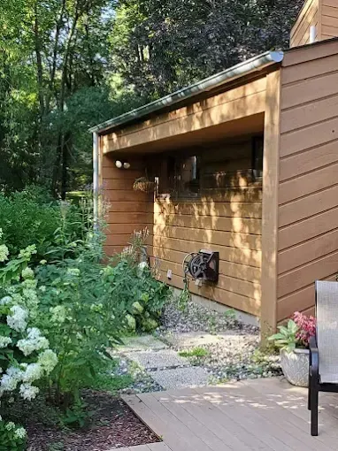 Brown building with covered patio and plants, with a gray walkway and woods in the background.