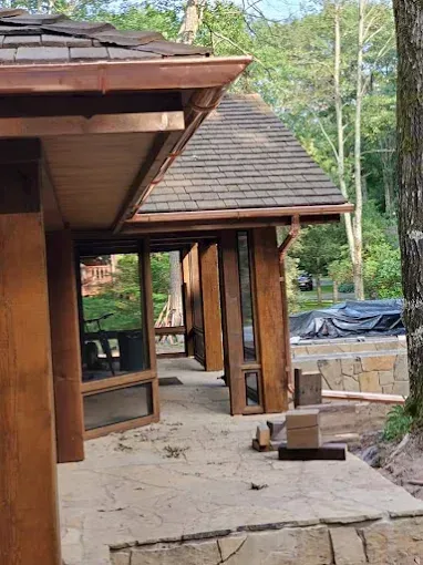 Wooden structure with a tiled roof and screened-in porch, built on a stone patio in a wooded area.