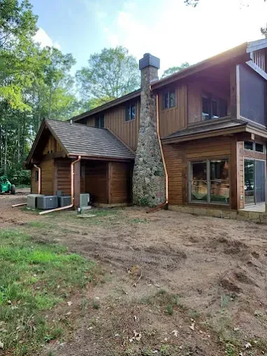 Two-story wood-sided house with stone chimney, brown roof, and surrounding dirt yard and trees.