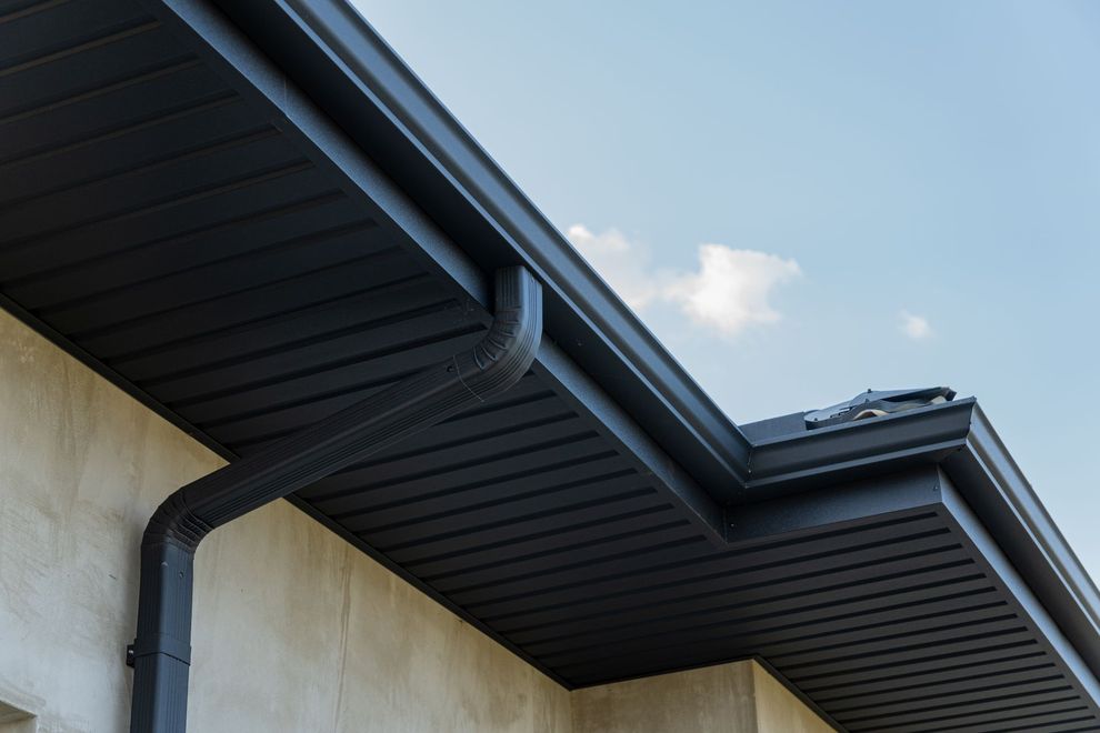 Black rain gutter system on a building with a light beige wall and a blue sky in the background.