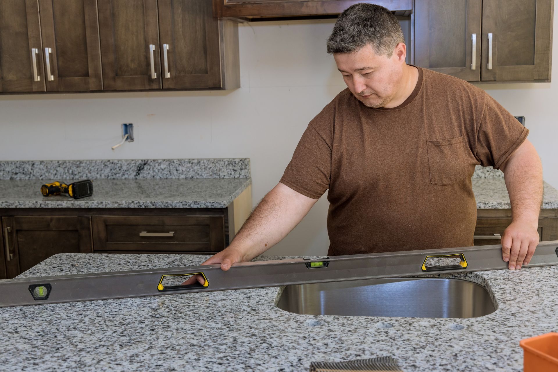 Man using a level to check countertop in a kitchen renovation. Dark cabinets, granite counters, tools visible.