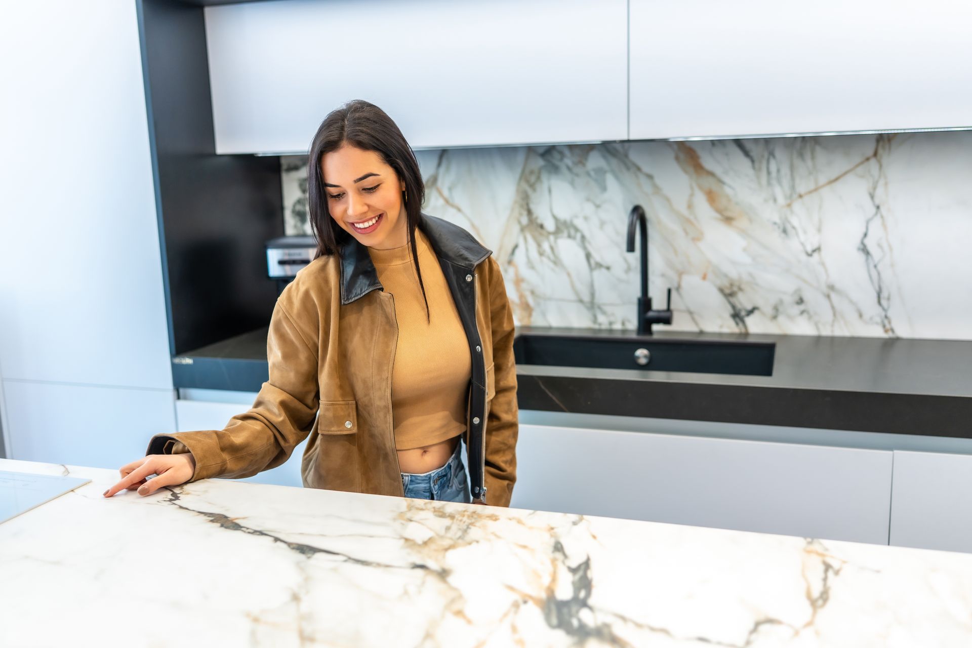 Woman smiles, touches countertop in modern kitchen with marble backsplash and black faucet.