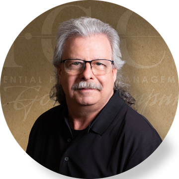 Man with graying hair and mustache wearing glasses, black shirt, against a brown background.