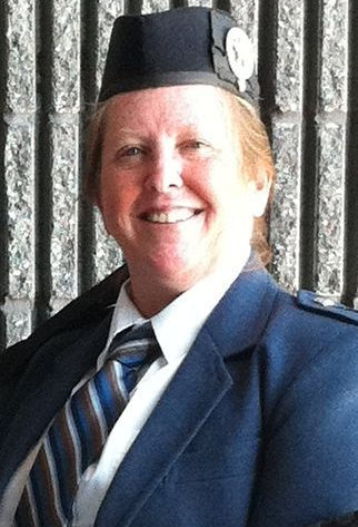 Woman in blue jacket, tie, and black hat smiles at camera. Against a vertical stripe background.