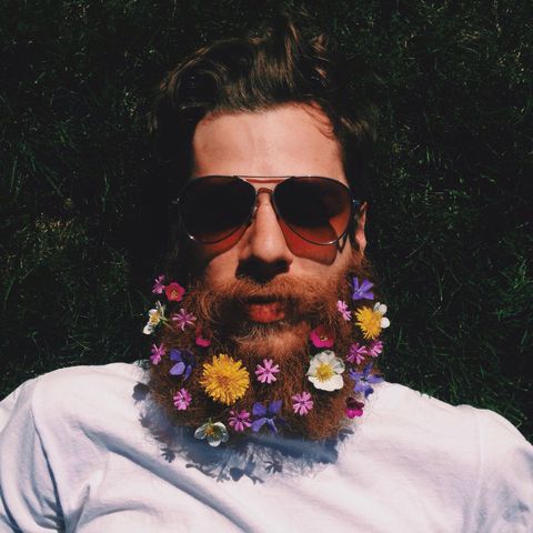 Man with beard decorated with colorful flowers, wearing sunglasses, lying on grass.