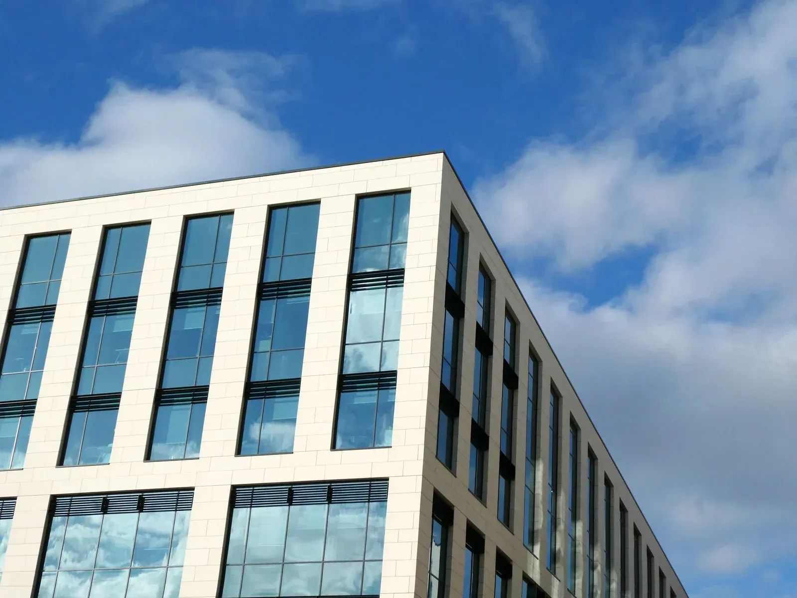 Modern office building with light stone facade and blue glass windows against a bright blue sky with puffy clouds.