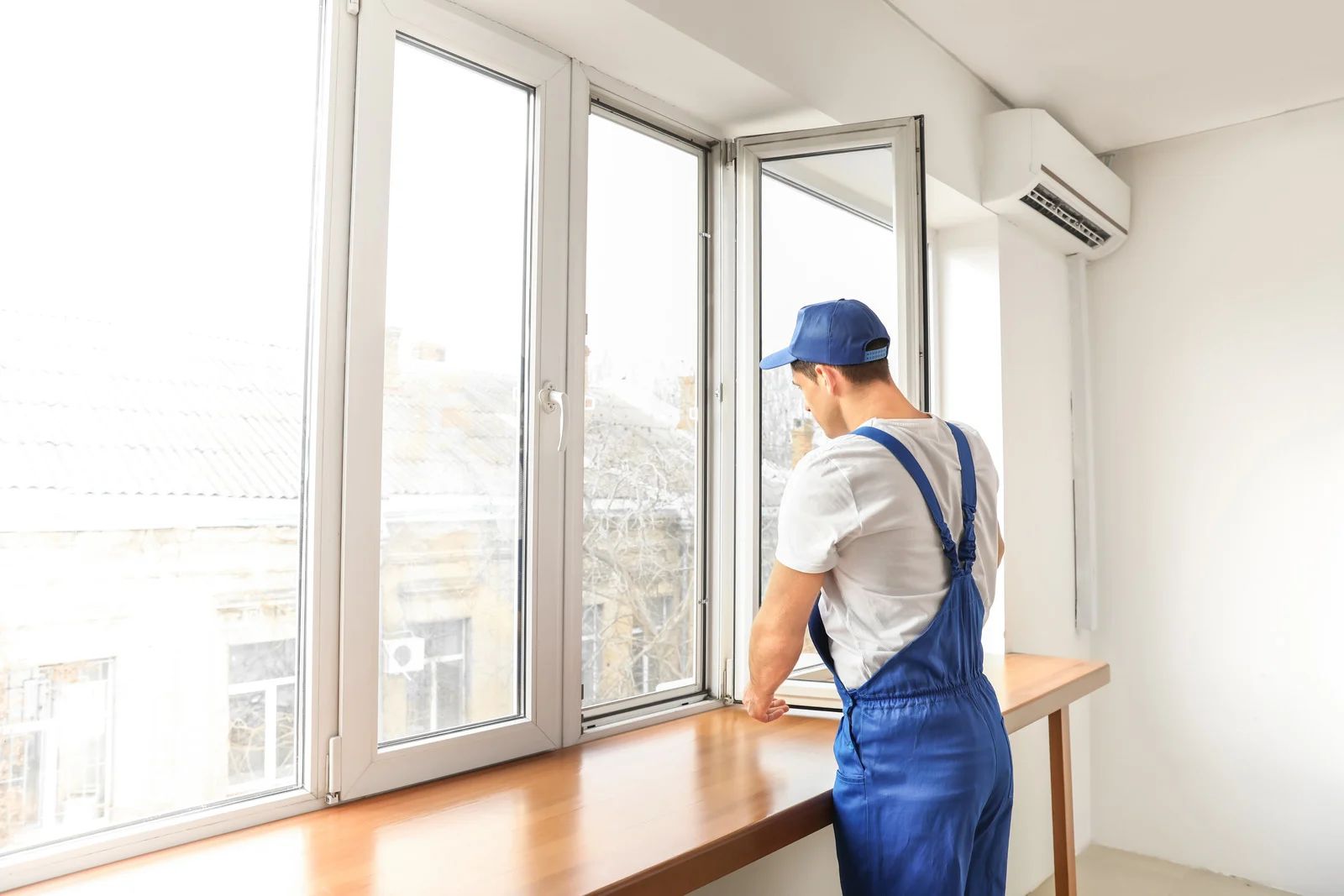 Man in blue overalls opening a large window in a bright room with a wooden ledge.