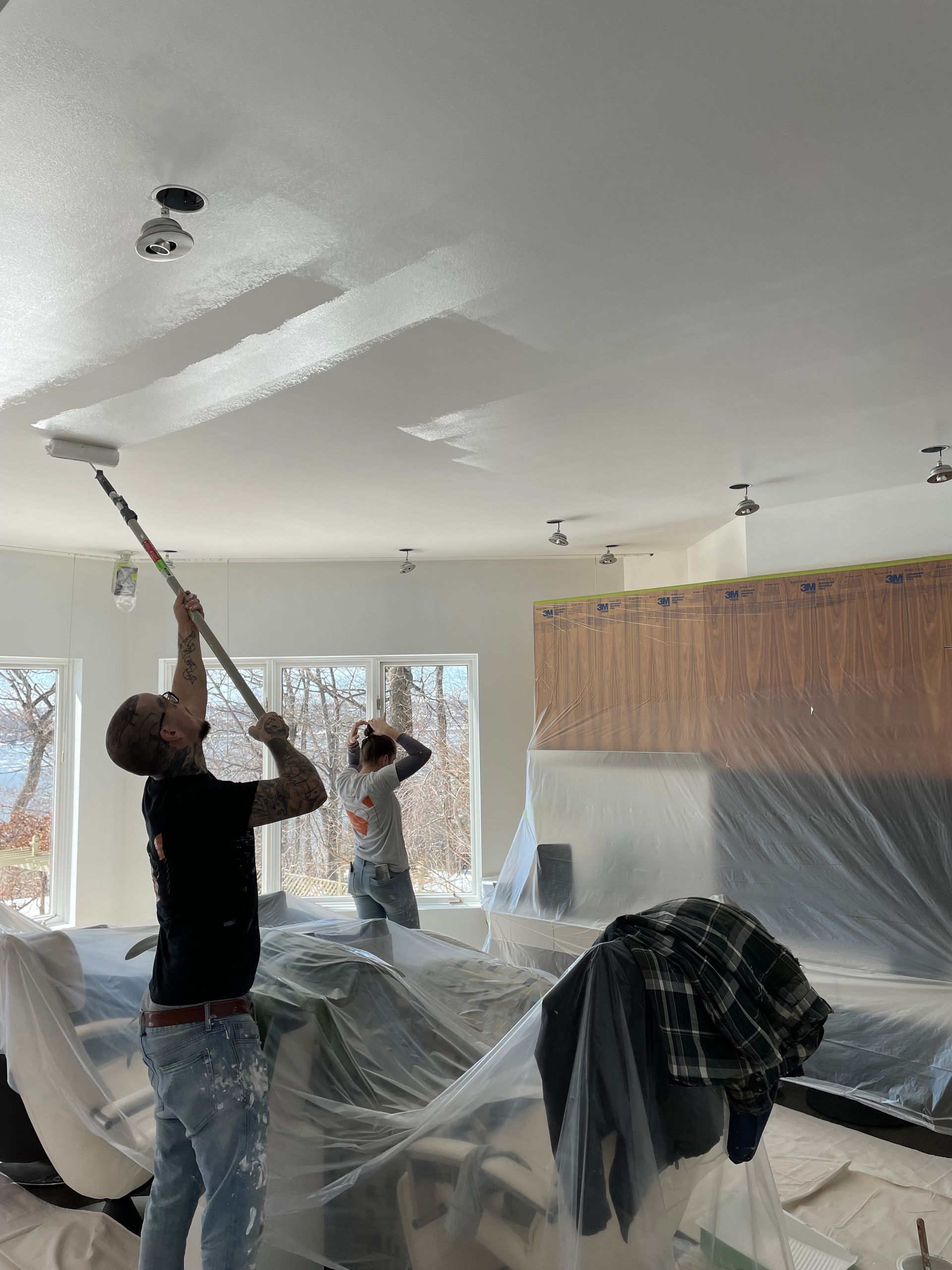 Deux personnes peignent un plafond blanc au rouleau ; intérieur d'une pièce, meubles recouverts de bâches en plastique.